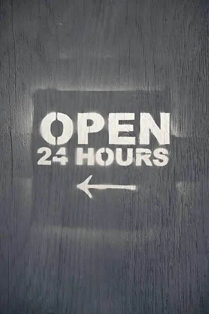 Open 24 Hours sign with arrow in stencil spraypaint on rough gray wood