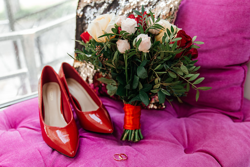 Red patent leather shoes of the bride near gold wedding rings and a bouquet of roses