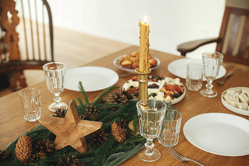 Christmas table setting. Stylish table runner with fir branches and pine cones, candles, cheese appetizers and starters, glasses and cutlery. Holiday dinner arrangement. Happy new year!