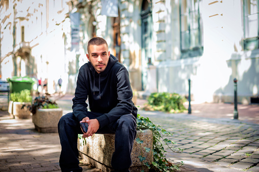 Close-up portrait of a relaxed young man sitting in a city street. Young male looking away and wearing hoodie.