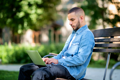 Young man using laptop for studying or working outdoor. Young male wearing casual clothes and sitting on the bench in the public park.