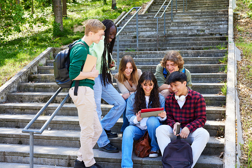 Teenage girl explaining homework over digital tablet to diverse friends while sitting on staircase at campus after school