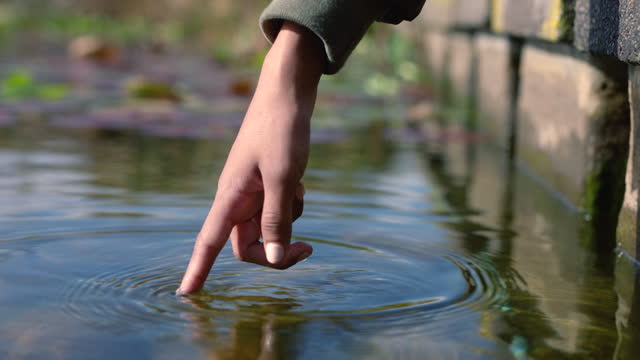 Close up woman's finger gently touching the surface of the water in a forest river or lake and uses her finger to make a circular movement causing spirals in the water outside. Patterns in a pond