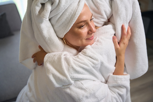 Loving mother tightly hugging her little pretty daughter, girls resting and relaxing in soft bathrobes and towels on heads after spa