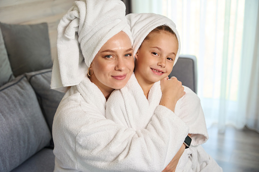 Beautiful mother and pretty little daughter in white soft bathrobes and bath towels on heads hugging and posing looking at camera