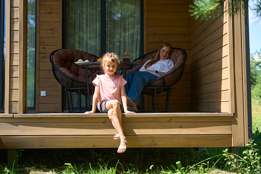 Attractive mother sitting in cozy garden armchair on wooden porch of country house and watching her little daughter playing nearby in grass, enjoying fresh air