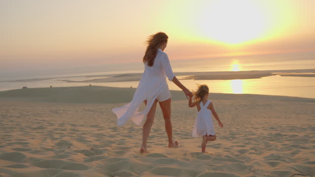 Mom and daughter in white clothes run barefoot along the sand holding hands at sunset on the ocean shore. Kid dream concept. happy family little girl and mom alone with nature relax sunset concept