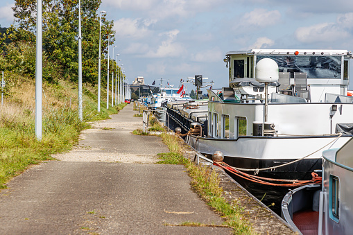 Pedestrian path along canal with cargo ship anchored at pier on coast, lane lock, street lamps post in line, green trees, sunny day with hazy blue sky in Ternaaien, Belgium