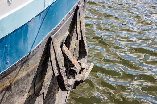 Closeup of mooring anchor on side of cargo ship, dirty and rusty, combination of white, blue and black colors, reflection of sunlight on water surface with wavy texture in background, sunny day