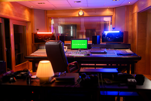 Photo of an interior of a music studio with a large mixer console with monitors and speakers and other studio equipment. In the back is a glass window of an isolation room silent booth for recording.
