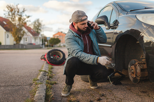 Young man talking on the phone while replacing his car tires outdoors.