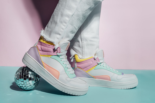 Close up photo of legs wearing retro style high-top multicolor sport sneakers shoes with disco ball on blue and pink background. Minimalism party, vintage retro style of 80s - 90s vibes