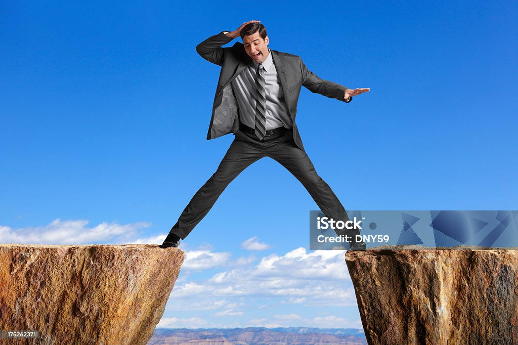 Businessman straddling the divide created by two cliffs A businessman straddling the divide created by two cliffs. At The Edge Of Stock Photo