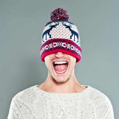 Portrait of a playful young adult man laughing at the camera with a winter cap covering his eyes. Studio shot on the grey background. 