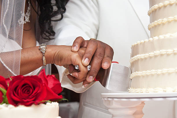 African American couple cutting white wedding cake with red roses A close up shot of a African American couple bride and groom cutting a white wedding cake on white table. Couple is wearing wedding rings and jewelry. Cake has ornate frosting. Red roses on top of tier cake. white wedding cake red roses stock pictures, royalty-free photos & images
