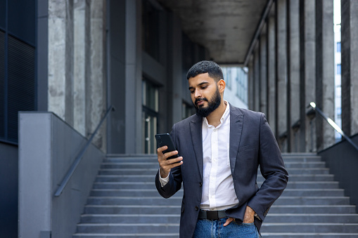 Young Indian man, real estate agent, broker standing near office outside and using phone, waiting for appointment, typing message.