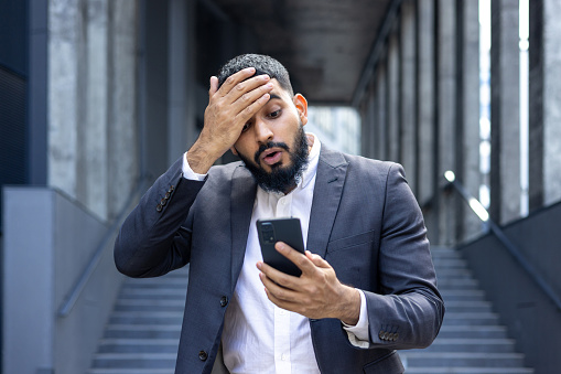 Shocked moody muslim male businessman standing near office building and looking upset at phone screen, holding head.