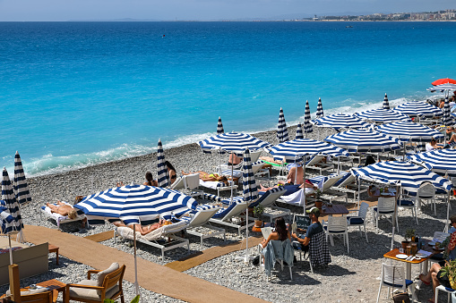 Nice, France - April 26, 2023: Blue-white umbrellas and sunbeds on a pebble beach for sunbathers to relax in both the shade and the sunlight here on the beach of the Promenade des Anglais.