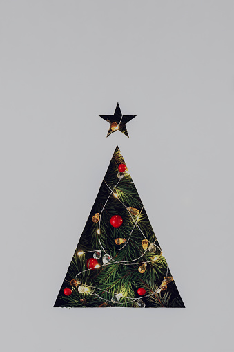 Triangular Christmas tree cut out in gray cardboard with lighted garland and Christmas tree branches.