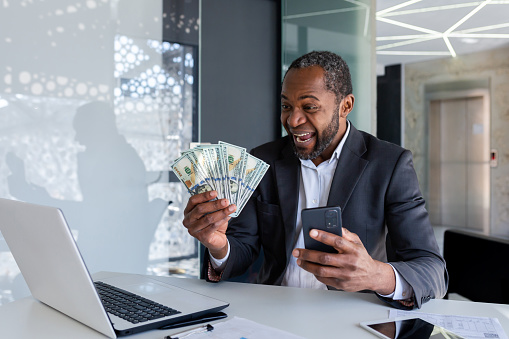 Happy African-American man sitting at the desk in the office, holding a mobile phone in his hand and looking enthusiastically at a fan of cash money.