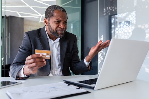 Upset and worried businessman at workplace, man cheated received online money transfer error, african american man unhappy sitting with laptop inside office holding bank credit card.