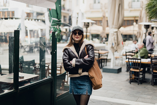 Portrait of a beautiful young fashionable woman wearing a cap and shearling jacket in the city street on a lovely autumn day in Thessaloniki
