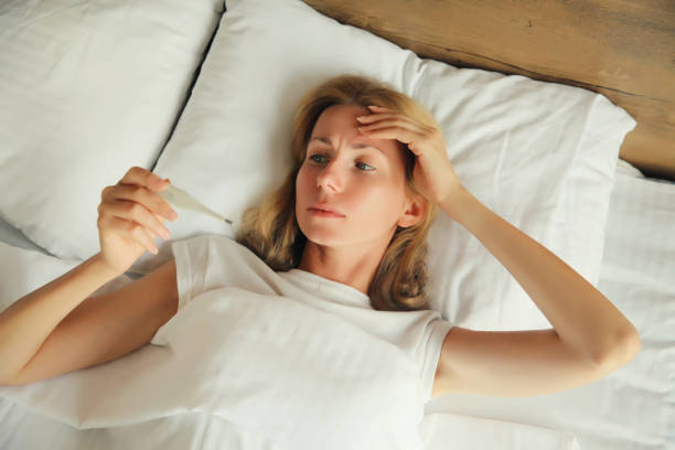 Sick woman measuring checking body temperature looking at thermometer while lying on bed at home stock photo