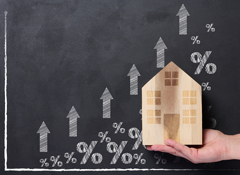 A hand holds a wooden house against the background of a graph with growing indicators, the concept of rising house prices, high rent and an increase in mortgage interest rates