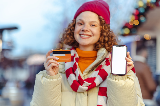 Front view on a young woman in warm clothing holding a smart phone with a white screen and credit card. She is looking at the camera