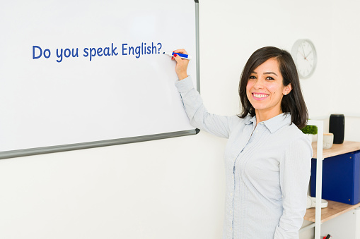 Beautiful English teacher smiling looking happy teaching a language lesson to the class while writing on the whiteboard