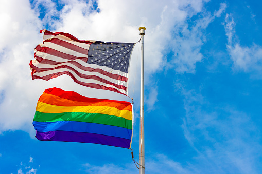 The LGBTQ rainbow flag flying underneath the American flag with the flagpole centered and copy space to the right side of the image.