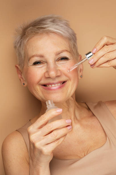 Happy senior lady applies cosmetic oil serum on face takes care of skin and smiles broadly enjoys beauty treatments stock photo