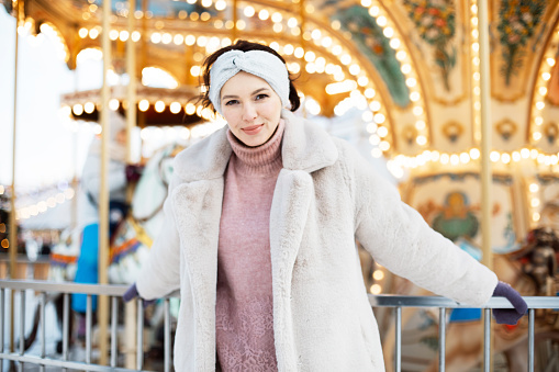 A beautiful young woman in a faux fur coat and snake clothes poses next to a carousel at a Christmas market.