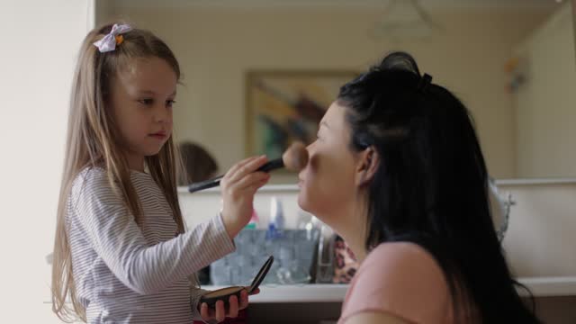 Pretty cute little girl doing make up for her mother with eye shadows palette, brush. Quality family time, enjoying domestic life, motherhood.