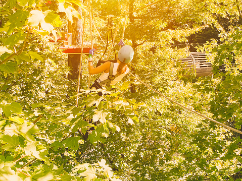 High-altitude climbing training of child on adventure track, equipped with safety straps and protective helmet.A teenager Girl walks along a rope bridge between trees in an amusement park in safety gear and a helmet. Rope park in wood forest.
