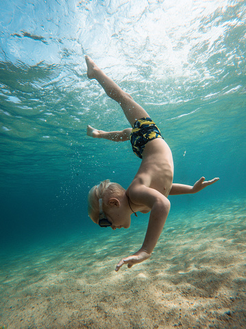 Small caucasian kid exploring underwater in turquoise color sea. He is wearing swimming goggles and swimwear. Trani Ammouda, Greece.