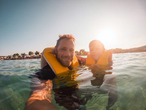 Young Caucasian father and his boy taking a photo in the water while enjoying a beautiful summer day at the beach. They are wearing yellow life jackets. Trani Ammouda, Greece.