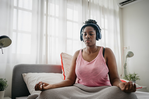 A black woman is sitting with closed eyes and listening to a meditation podcast at home.