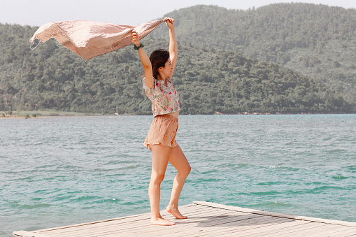 This is an image of a young Caucasian, European woman standing on a wooden pier beside the blue sea and green mountains and hills on a bright sunny summer day. She is also holding a brown large towel floating in the wind and her body is elegantly posed.
