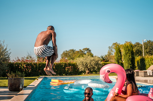 A cheerful black male adds to the fun at a pool party by performing a cannonball jump, creating a lively splash, and sharing laughter with diverse friends.