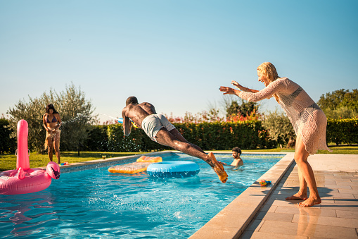 A carefree moment at a pool party as a Caucasian female pushes a black male into the pool, surrounded by their diverse group of friends, all sharing in the laughter and joy.