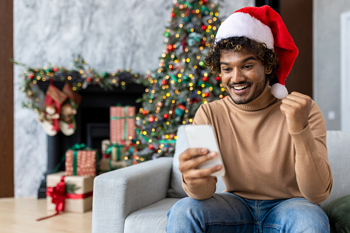 Man in red holiday santa hat sitting on christmas in living room on sofa near tree, hispanic man celebrating victory successful achievement and triumph, using app on smartphone, happy winner.