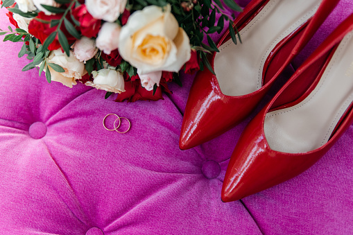 Red patent leather shoes of the bride near gold wedding rings and a bouquet of roses, top view
