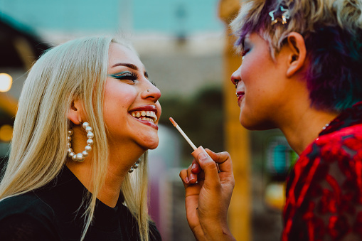 Young woman smiling as her friend applying lipstick on her. Friendship and beauty concept.