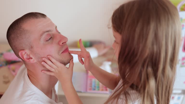 Child girl putting on make up on father's face , using decorative cosmetics, have fun together, spend time at home