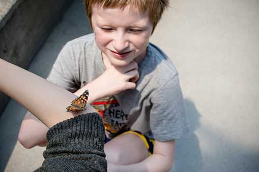 A candid and heartwarming lifestyle moment unfolds as a Painted Lady butterfly lands on a teenager's arm, while a 10-year-old looks on with a subtle smile of fascination, capturing the genuine connection between youth and nature