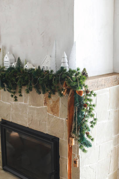 istock Modern christmas trees, houses, pine cones and spruce branches on fireplace mantel with brass bells and ribbon. Stylish decorated christmas fireplace in minimal scandinavian living room 1753384011
