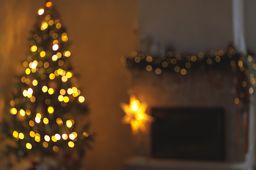 Christmas background golden bokeh. Atmospheric christmas eve at fireplace. Blurred image of illuminated christmas tree and festive decorated fireplace with lights in evening scandinavian room
