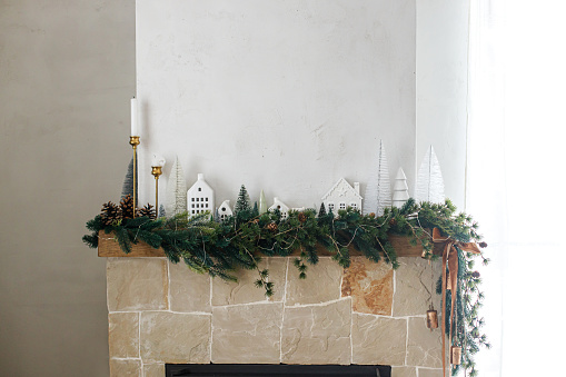 Stylish decorated christmas fireplace. Modern christmas trees, houses, pine cones and spruce branches on fireplace mantel with brass bells and ribbon. Christmas scandinavian living room decor