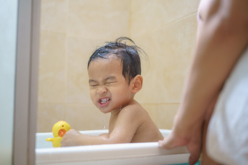 Adorable Asian little boy playing with bath toy in bathtub while his mother bathing him.